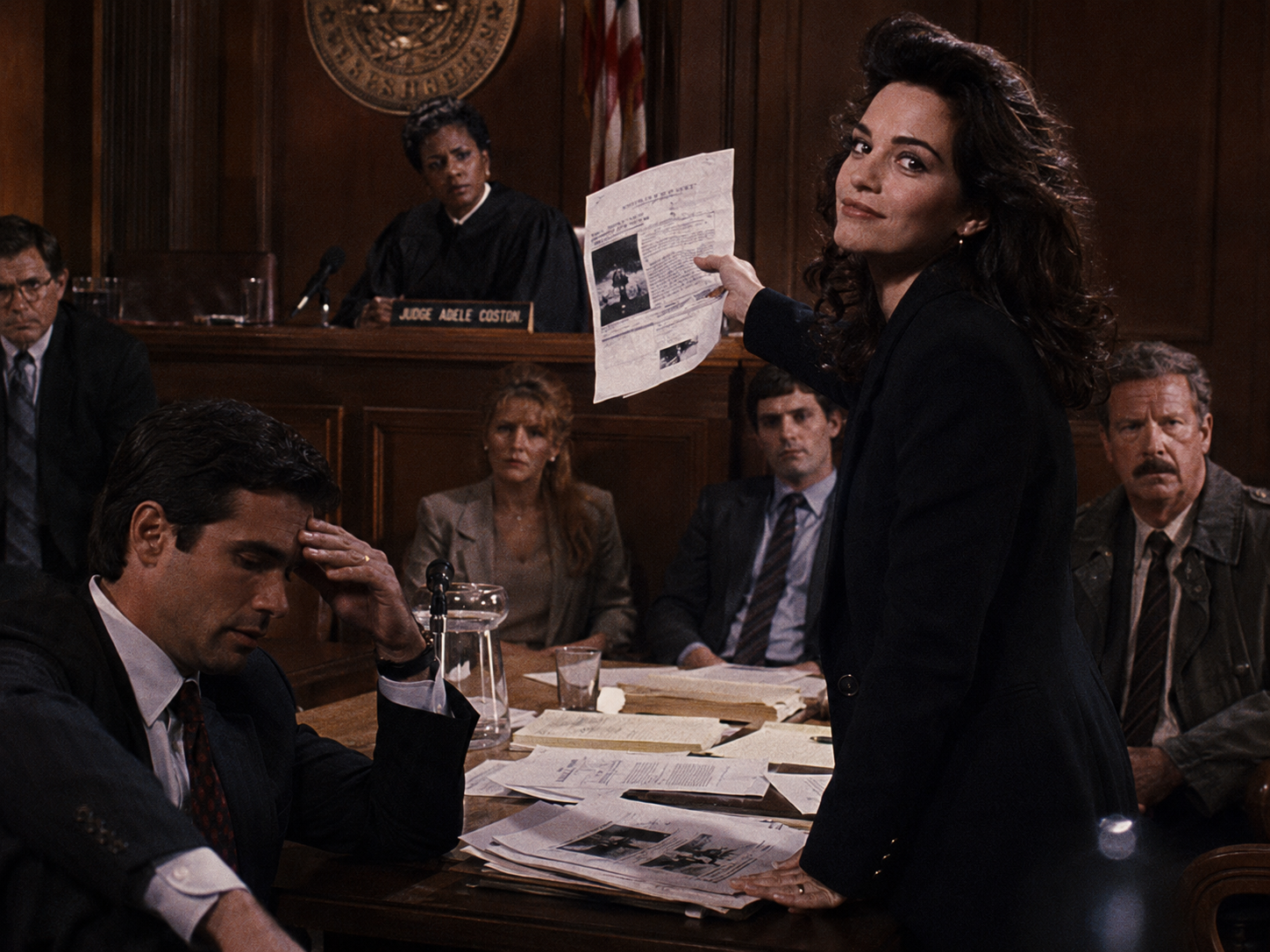 Nora brandishes a document in court as Stiles slumps